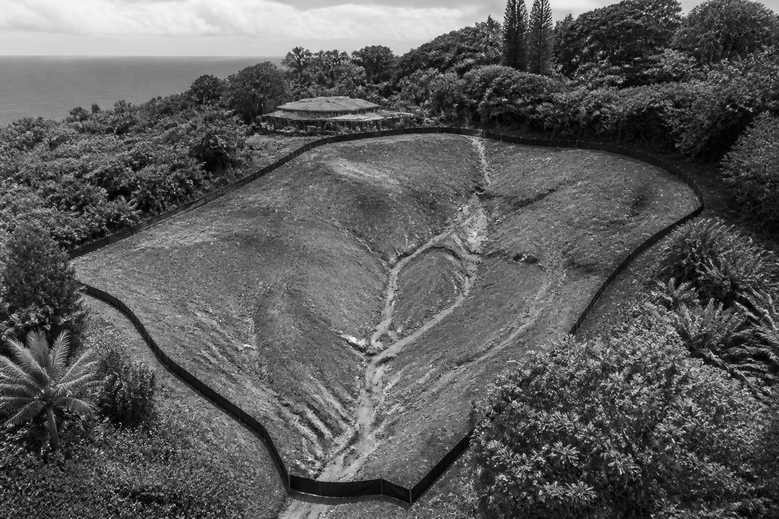 Aerial black and white photo of a cleared Kauaʻi hillside parcel with visible runoff erosion channels through exposed soil, silt fencing around the perimeter